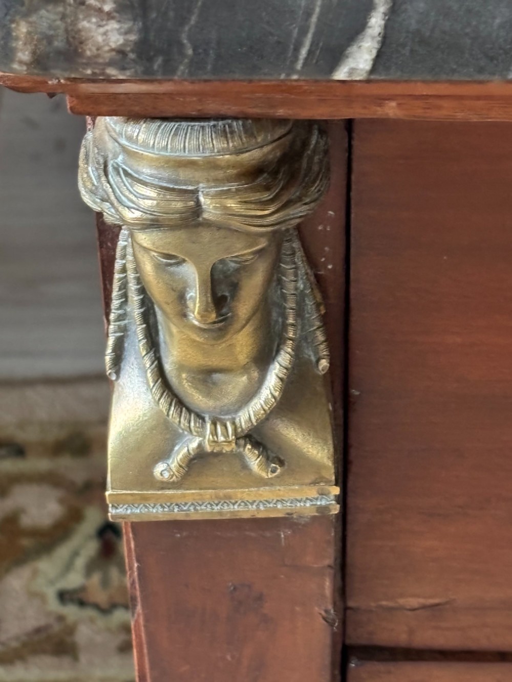 An early nineteenth-century French Empire mahogany commode featuring three drawers with brass lion-mask ring handles, gilt female caryatid mounts at the upper corners, a dark veined marble top, and sculpted paw feet. View VII