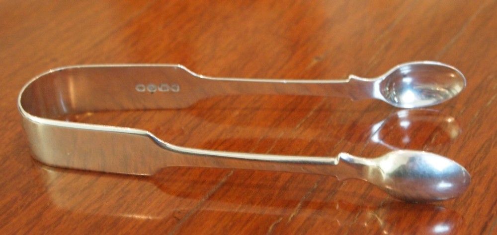 A pair of hallmarked antique sterling silver sugar tongs with spoon-shaped grips on a wooden background.