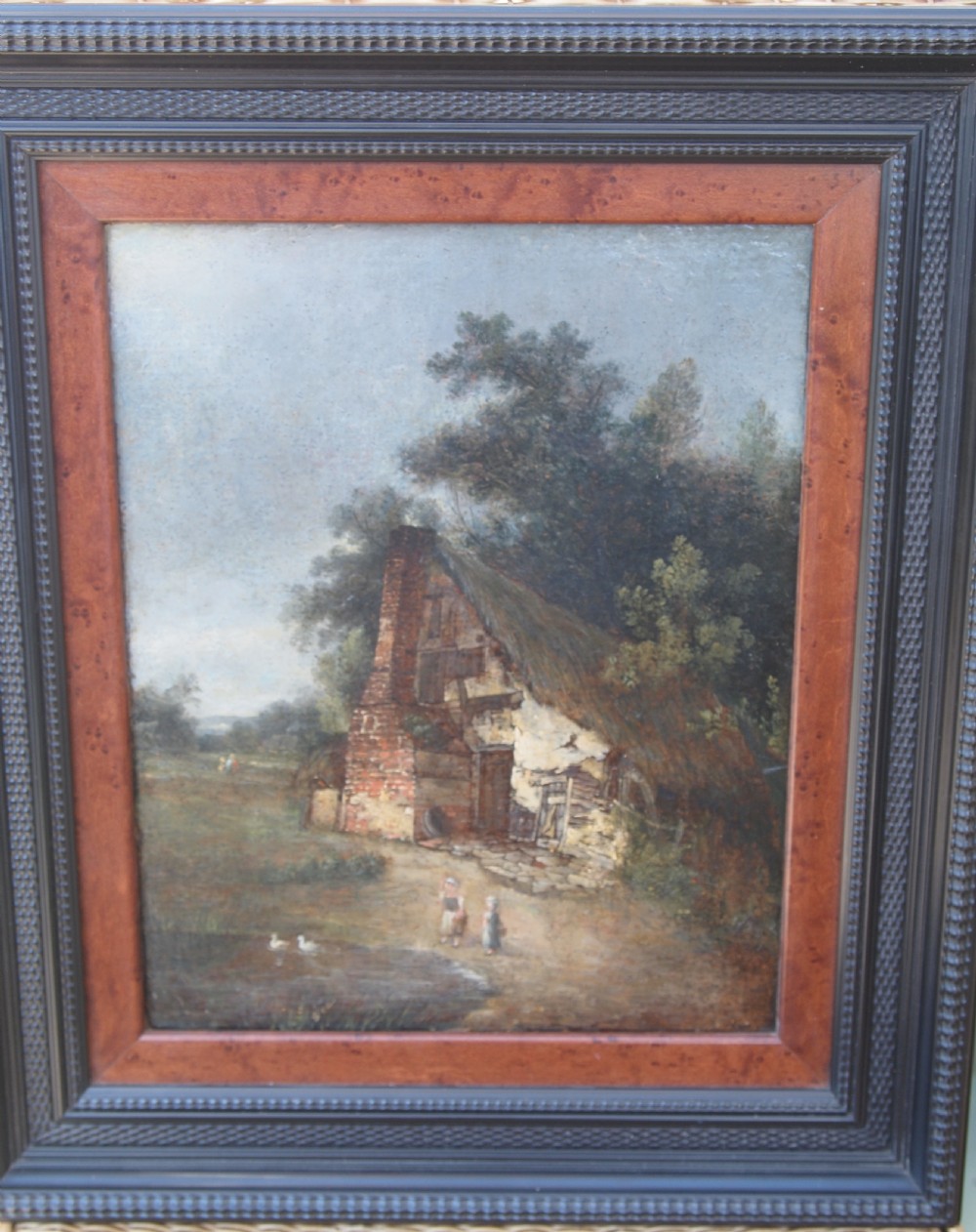A 19th-century oil painting depicting a thatched brick cottage nestled among trees with figures near a pond, housed in a moulded dark wood frame.