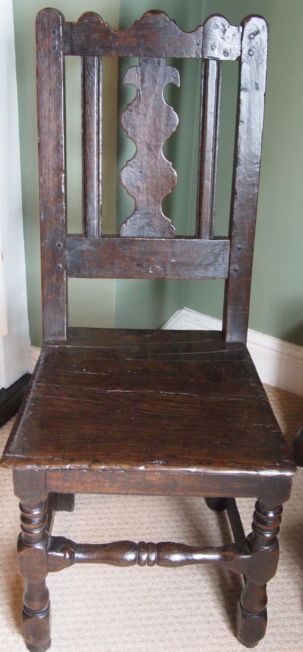 A 17th-century English dark oak side chair featuring a shaped crest rail, central carved splat, solid wood seat, and turned front legs.