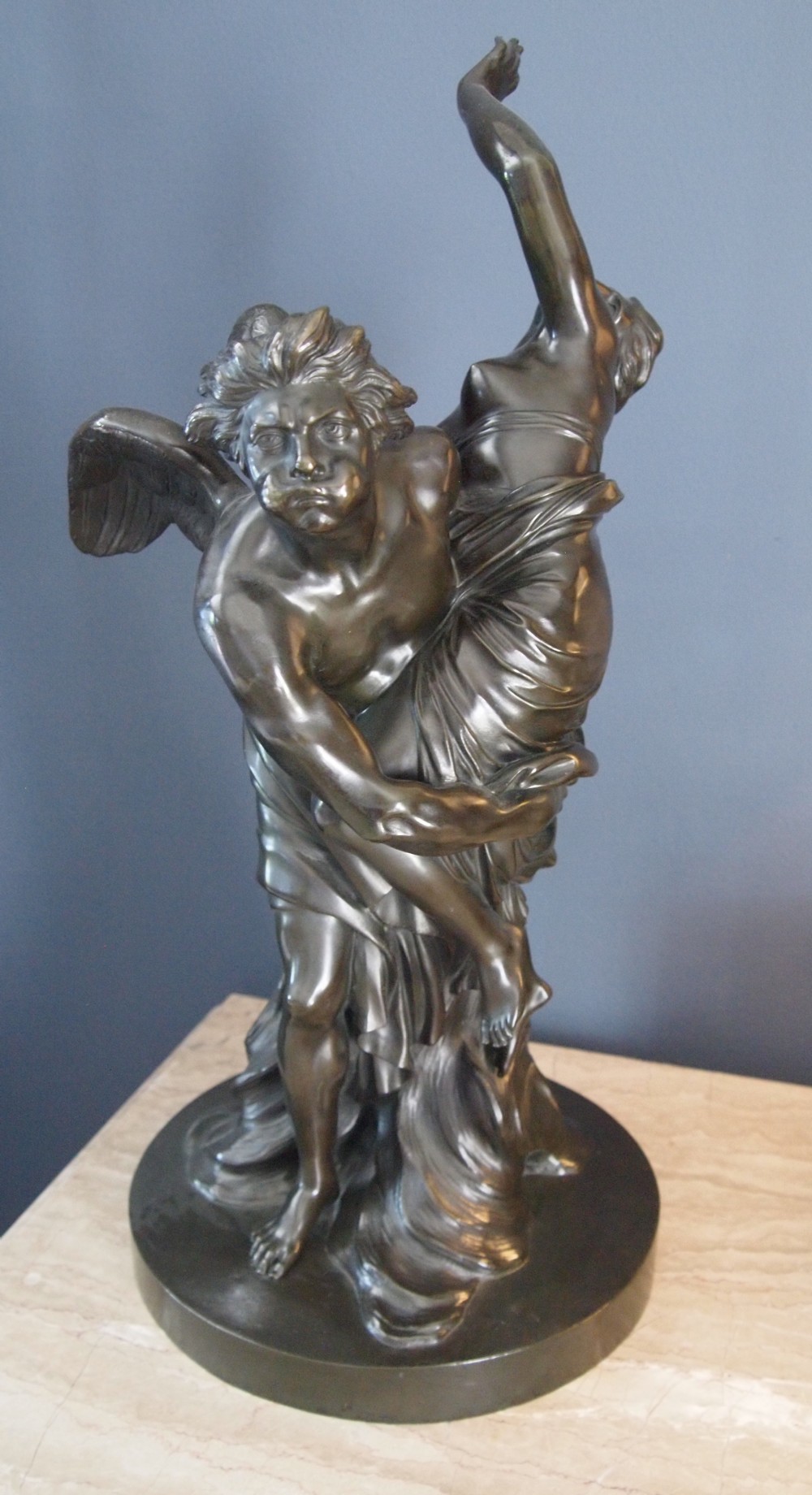 A nineteenth-century dark patinated bronze sculpture depicting a winged male classical figure carrying a draped female figure on a circular base.