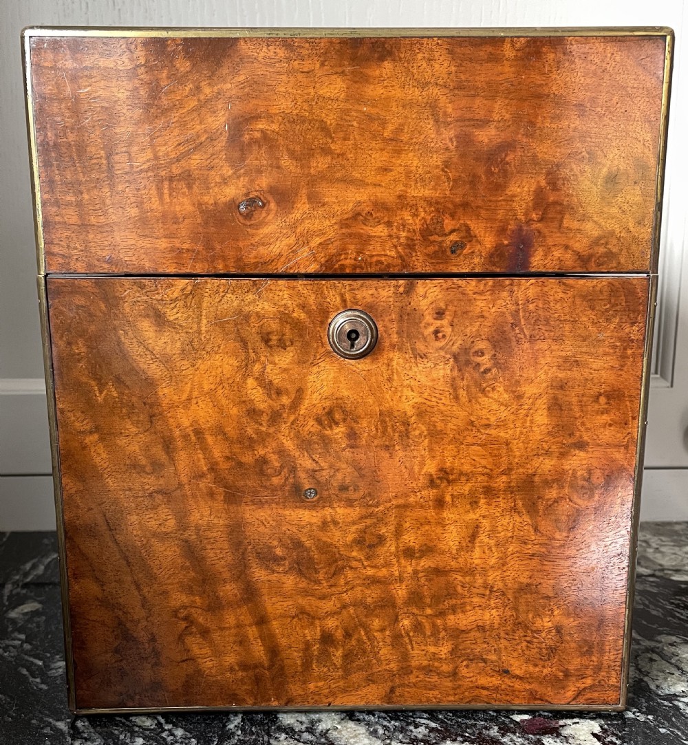 A closed rectangular burr walnut decanter box featuring thin brass trim around the outer edges and a circular brass keyhole escutcheon.