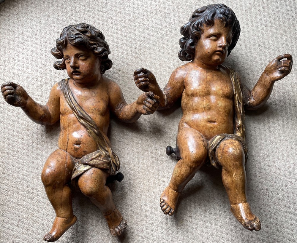 A pair of 17th-century French carved limewood putti featuring curly hair, draped sashes, and dynamic seated poses with a warm aged surface patina.