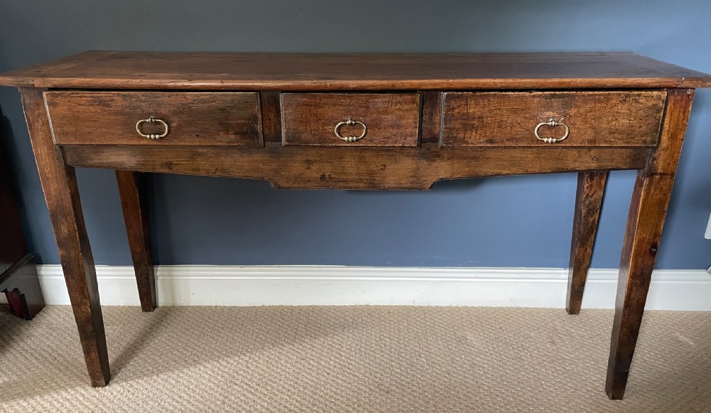 A nineteenth-century Chinese elm side table featuring three drawers with brass drop handles and straight legs set against a dark blue wall.