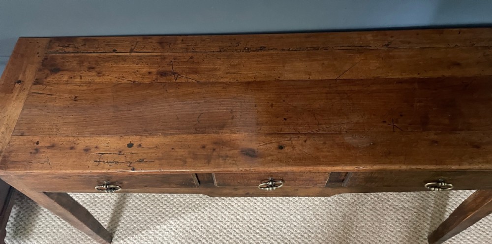 A nineteenth-century Chinese elm side table featuring three drawers with brass drop handles and straight legs set against a dark blue wall. View V