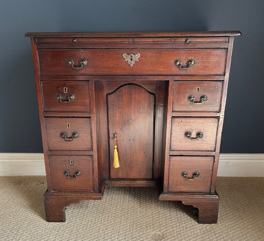An antique Georgian Cuban mahogany kneehole desk featuring six small pedestal drawers flanking a central recessed arched cupboard door, a full-width top drawer, brass handles, and bracket feet.