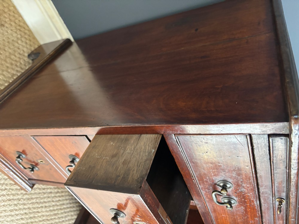 An antique Georgian Cuban mahogany kneehole desk featuring six small pedestal drawers flanking a central recessed arched cupboard door, a full-width top drawer, brass handles, and bracket feet. View III