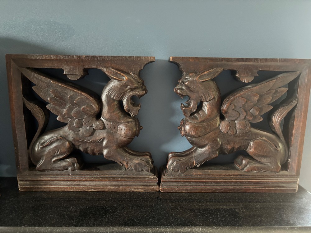 A facing pair of 19th-century carved dark oak panels depicting winged griffins set within rectangular timber frames.