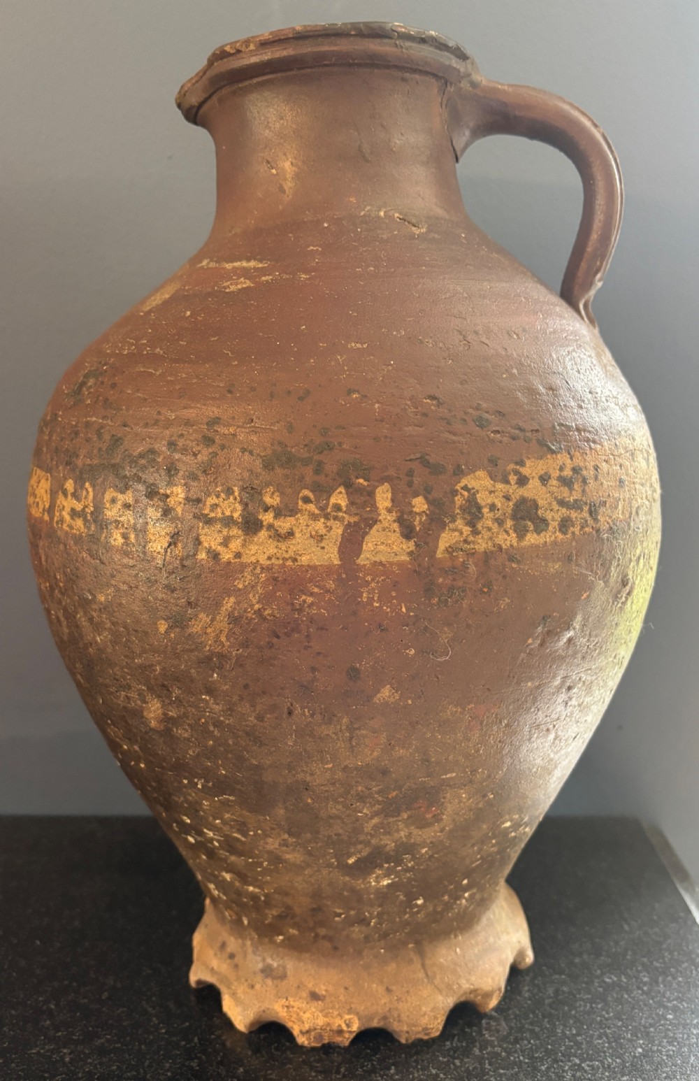 A large 17th-century earthenware tavern jug featuring a rounded body, piecrust footed rim, strap handle, and faded slip decoration.