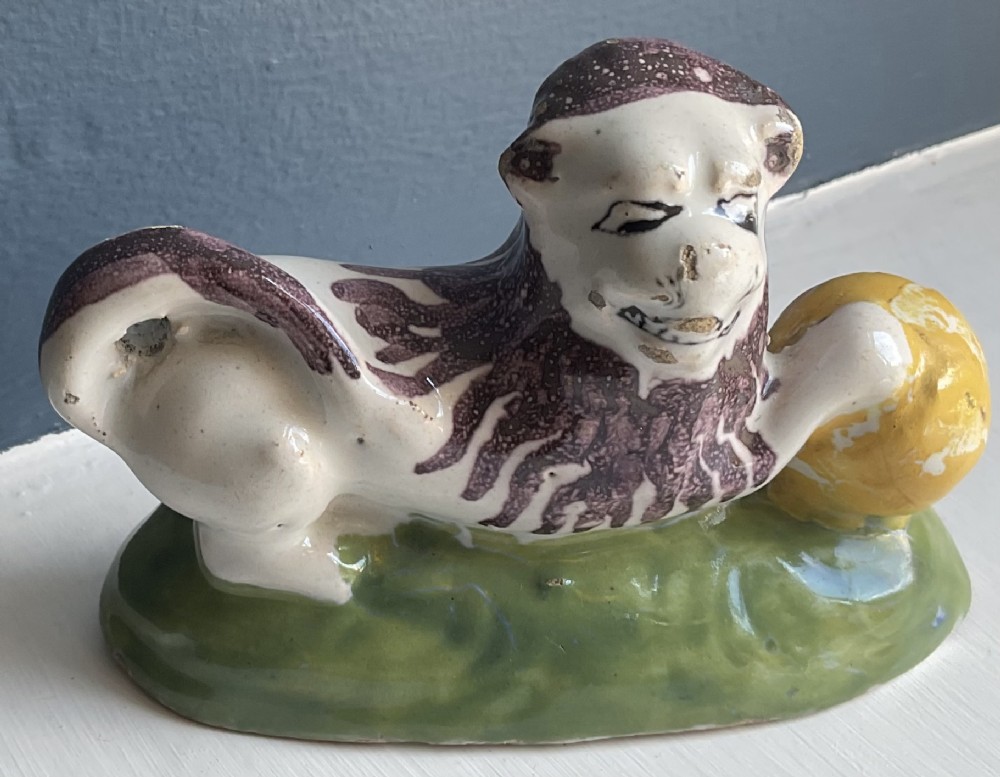 A tin-glazed ceramic figure of a reclining lion resting its paw on a yellow ball, featuring manganese brush-painted accents and a green base.