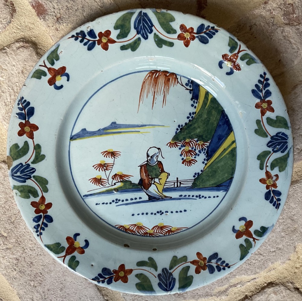 A circular tin-glazed earthenware delft plate featuring a central hand-painted figure in green, red, and yellow, surrounded by floral rim borders.