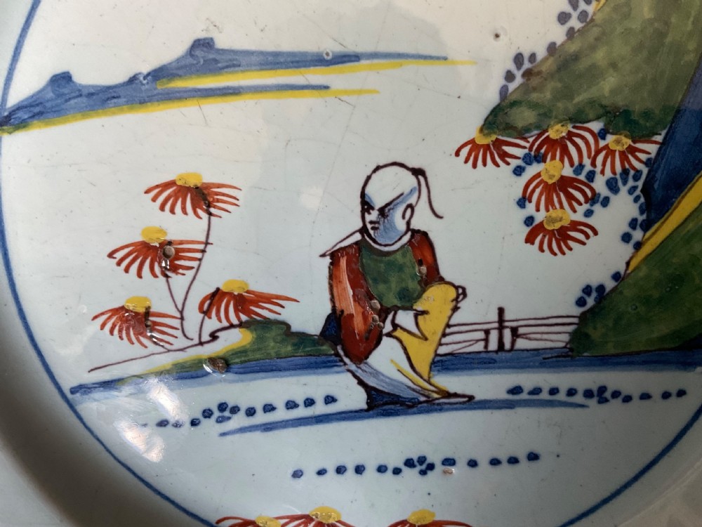 A circular tin-glazed earthenware delft plate featuring a central hand-painted figure in green, red, and yellow, surrounded by floral rim borders. View II