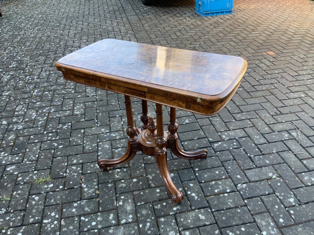 A Victorian burr walnut fold-top card table with highly figured wood grain, turned column supports, a central finial, and four carved splayed legs.