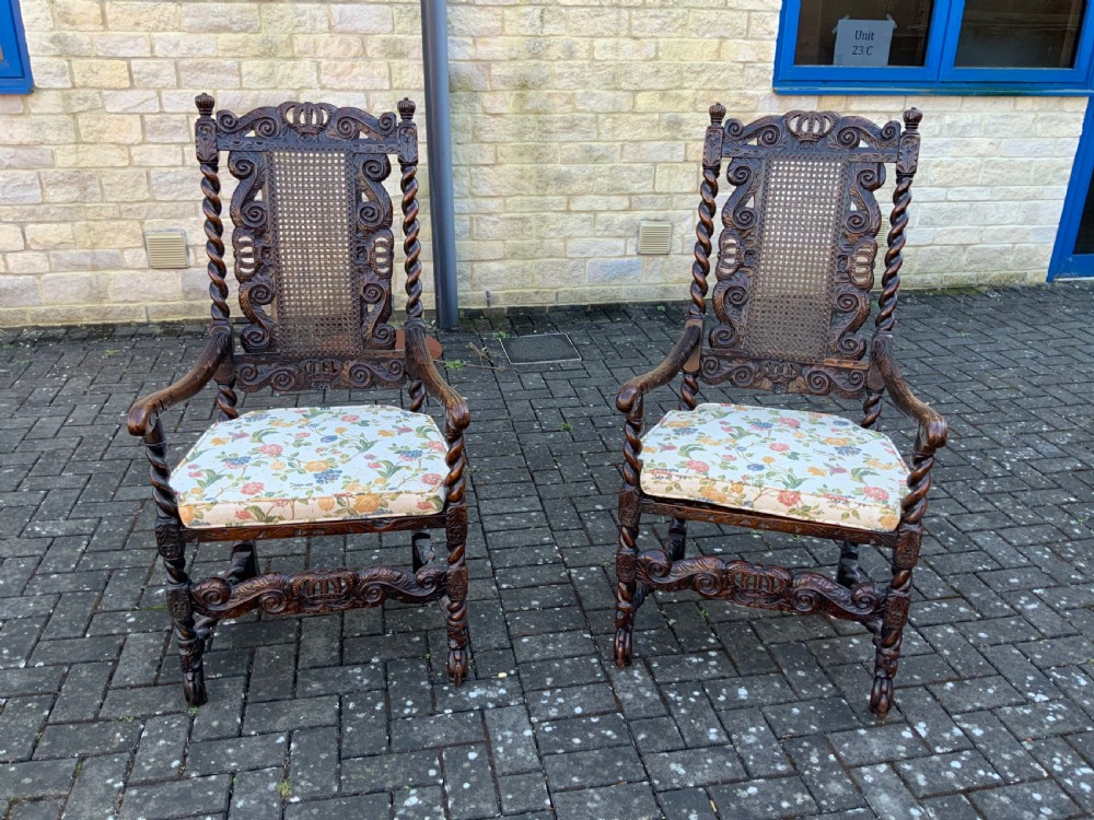 A pair of dark carved oak armchairs featuring high cane backs with carved crown crests, barley twist uprights, curved armrests, and floral upholstered seats.