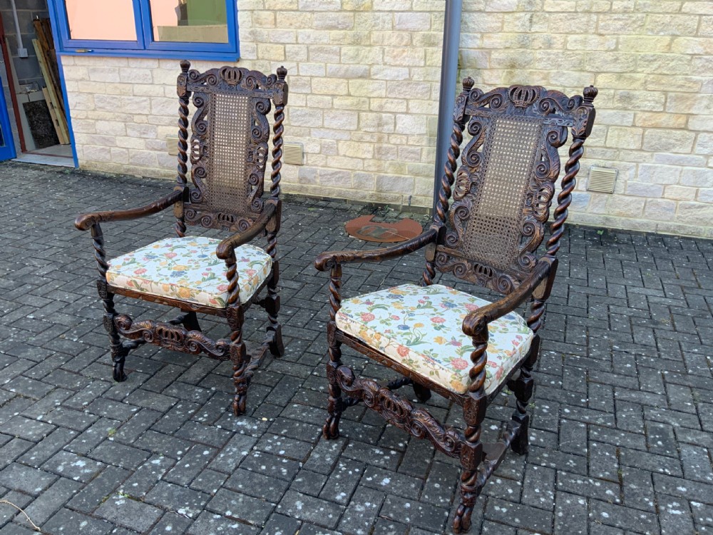 A pair of dark carved oak armchairs featuring high cane backs with carved crown crests, barley twist uprights, curved armrests, and floral upholstered seats. View V