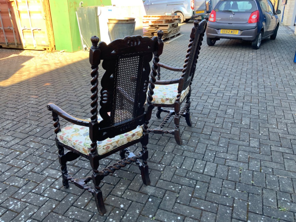 A pair of dark carved oak armchairs featuring high cane backs with carved crown crests, barley twist uprights, curved armrests, and floral upholstered seats. View VI