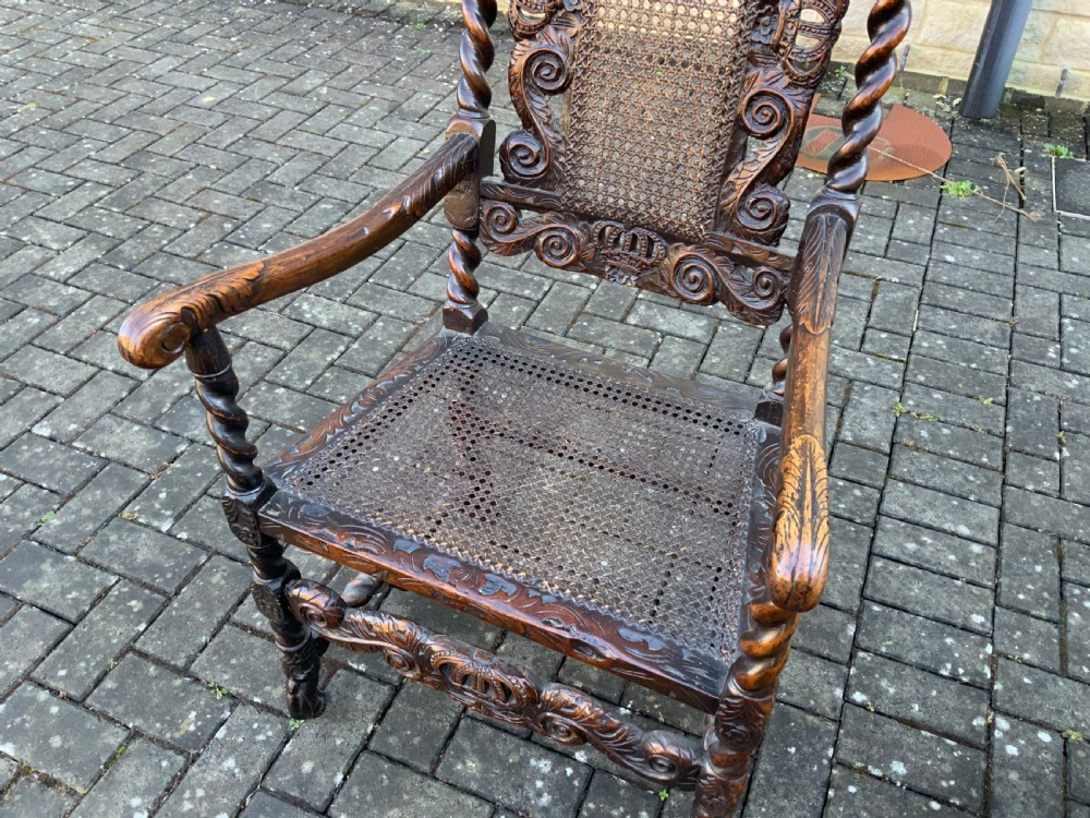 A pair of dark carved oak armchairs featuring high cane backs with carved crown crests, barley twist uprights, curved armrests, and floral upholstered seats. View X