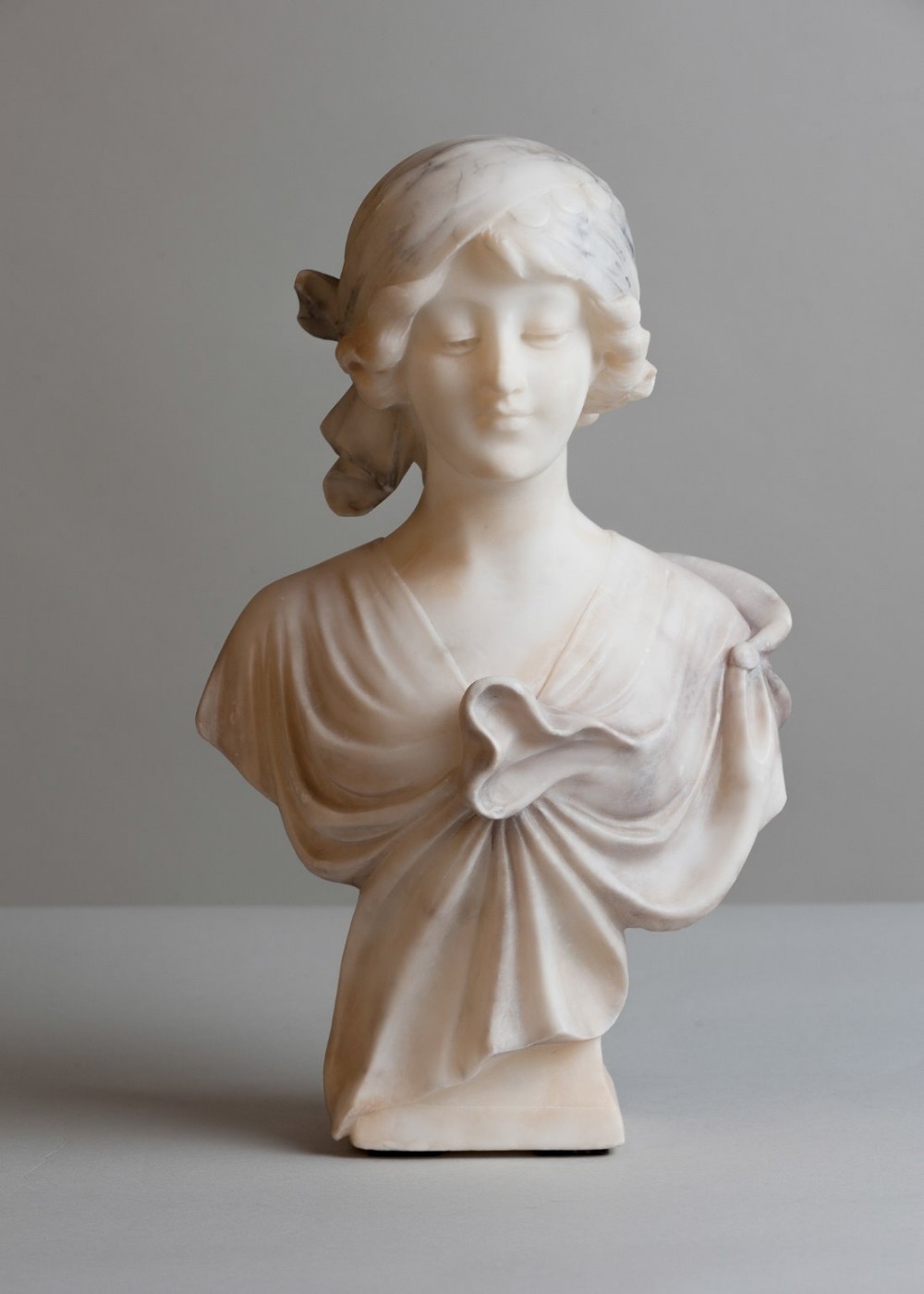 A carved cream alabaster bust depicting a young woman wearing a tied headscarf and draped gown, looking downwards.
