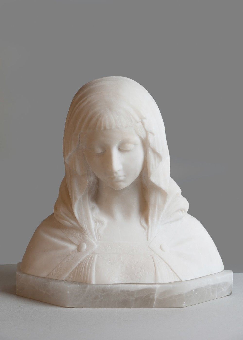 A carved white alabaster bust of a veiled female figure with downcast eyes resting on a multi-sided base.