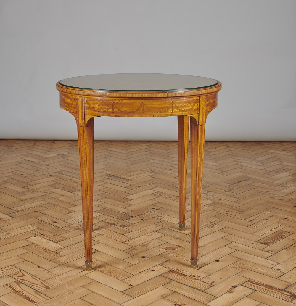 A circular French satinwood occasional table featuring inlaid floral swags along the apron, slender tapered legs with brass feet, and a fitted glass top.