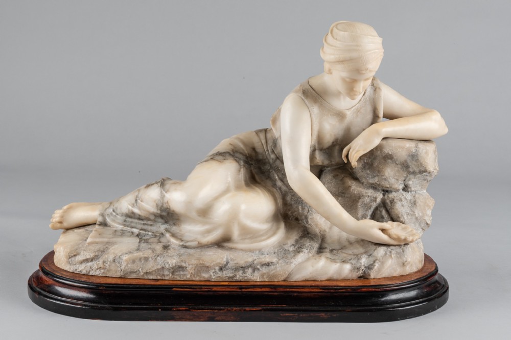 A carved two-tone alabaster sculpture depicting a reclining draped female figure resting on a rocky outcrop, mounted on an oval wooden base.