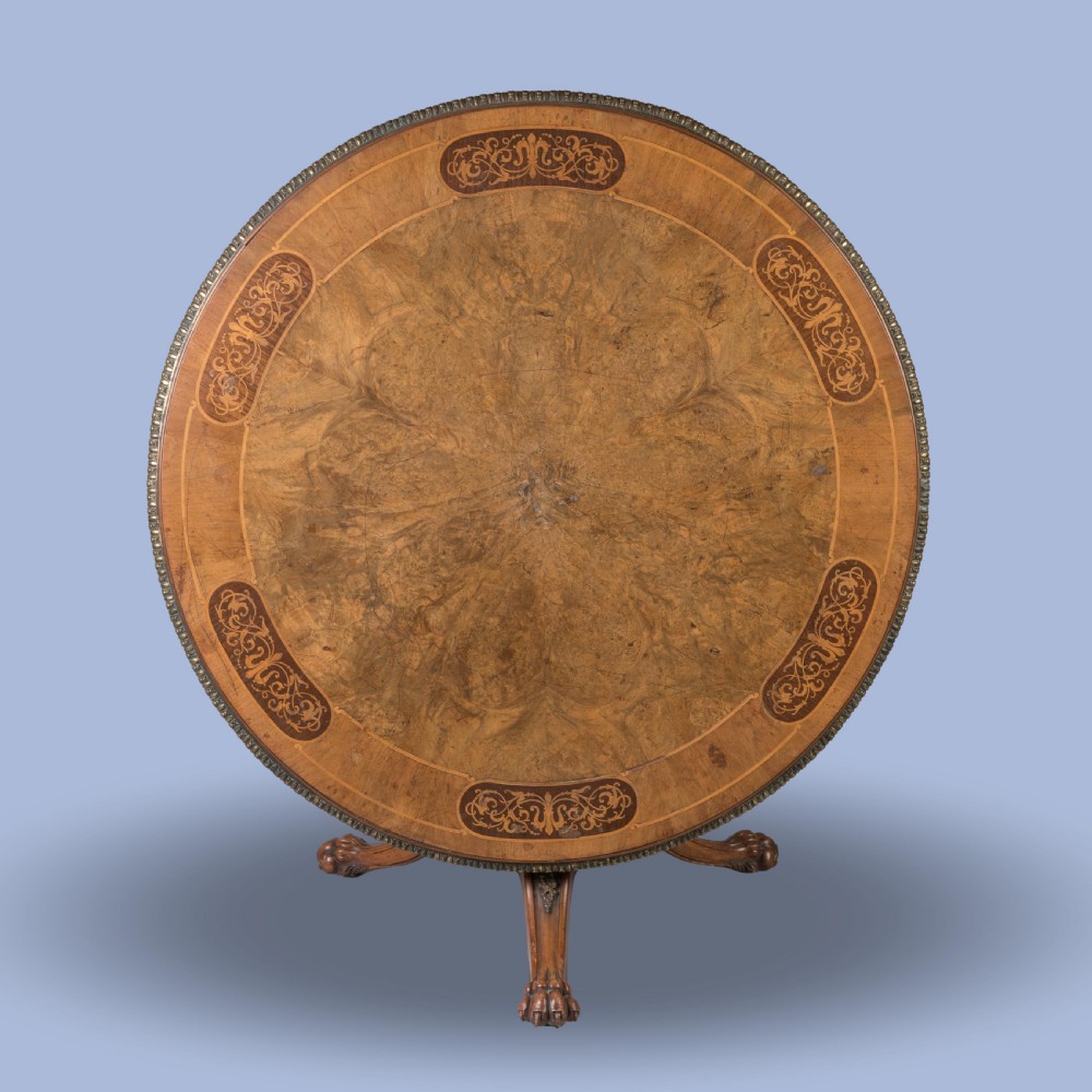 A circular Victorian burr walnut centre table featuring inlaid marquetry cartouches along the border and a carved pedestal base.