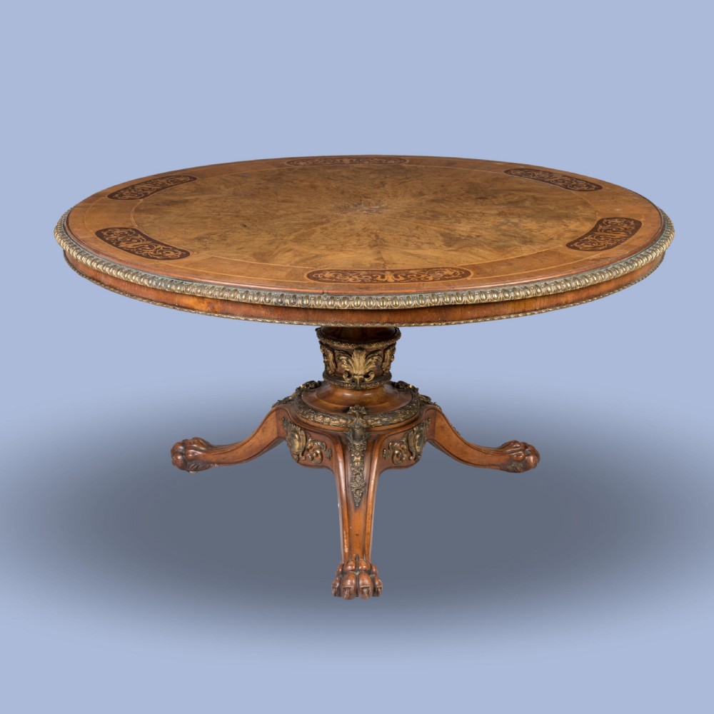 A circular Victorian burr walnut centre table featuring inlaid marquetry cartouches along the border and a carved pedestal base. View VI