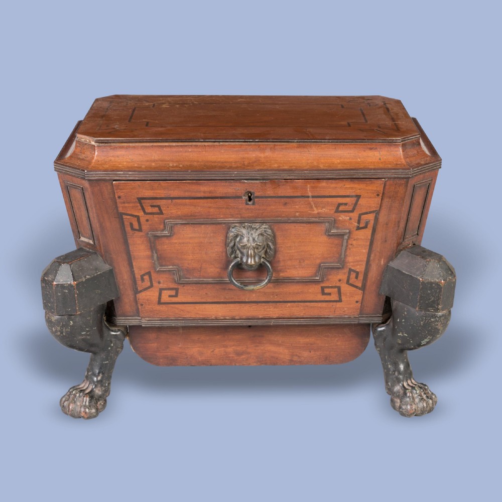 A Regency mahogany sarcophagus-form wine cellarette featuring Greek key inlay, a central brass lion-mask ring handle, and ebonised monopodia lion paw legs.