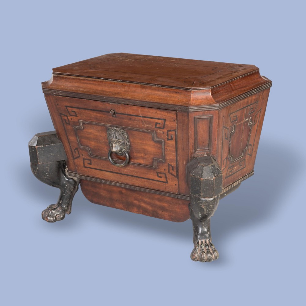 A Regency mahogany sarcophagus-form wine cellarette featuring Greek key inlay, a central brass lion-mask ring handle, and ebonised monopodia lion paw legs. View II