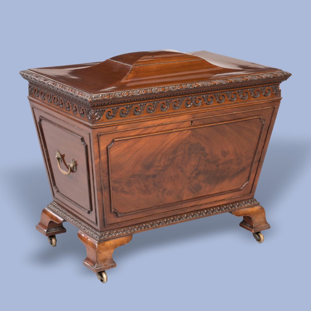 A sarcophagus-shaped antique mahogany wine cooler with carved decorative borders, side brass carrying handles, and brass castors.