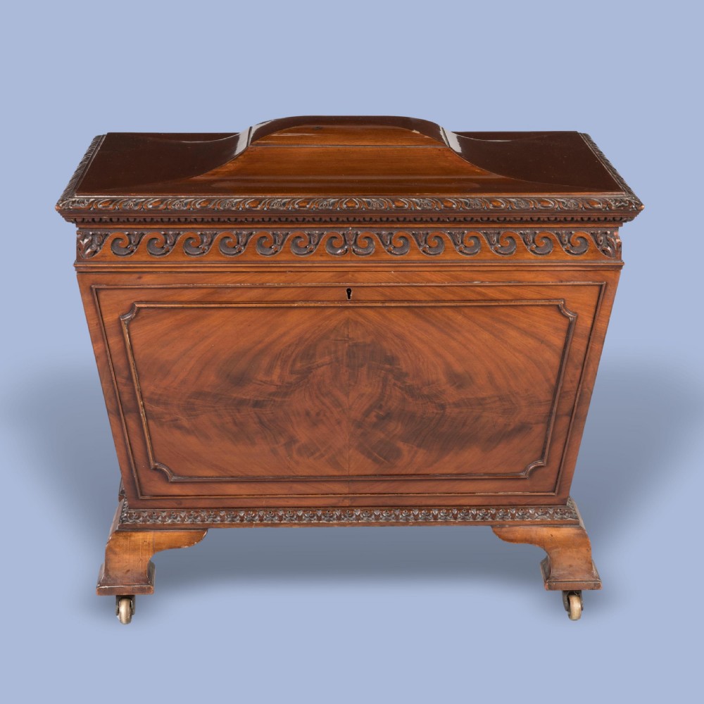 A sarcophagus-shaped antique mahogany wine cooler with carved decorative borders, side brass carrying handles, and brass castors. View II
