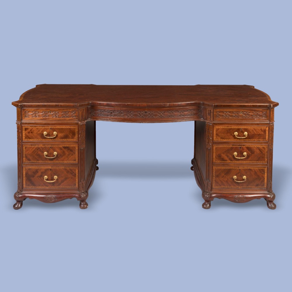 An antique mahogany pedestal desk featuring a serpentine front, parquetry drawer fronts, brass handles, and carved blind fretwork along the frieze.