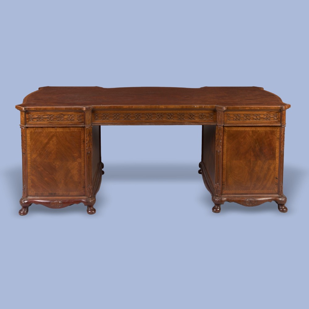 An antique mahogany pedestal desk featuring a serpentine front, parquetry drawer fronts, brass handles, and carved blind fretwork along the frieze. View II