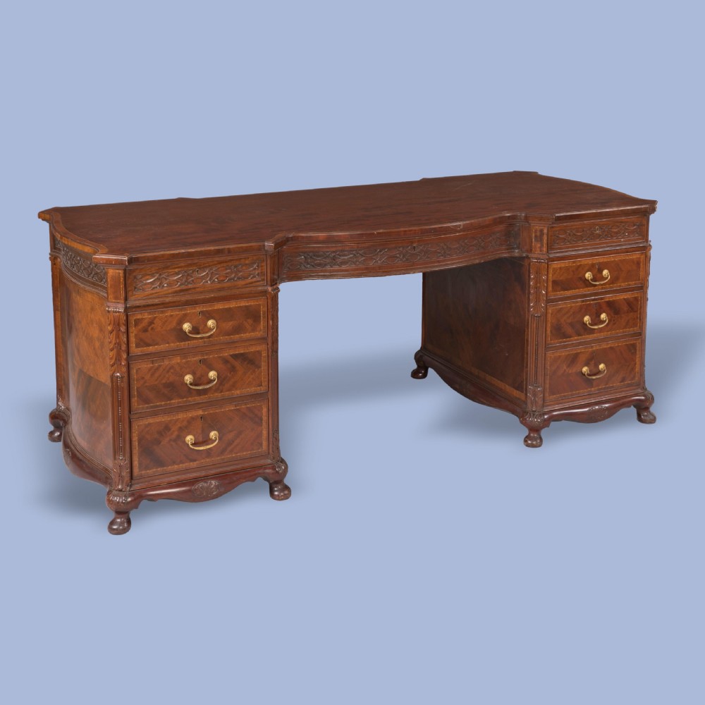 An antique mahogany pedestal desk featuring a serpentine front, parquetry drawer fronts, brass handles, and carved blind fretwork along the frieze. View III
