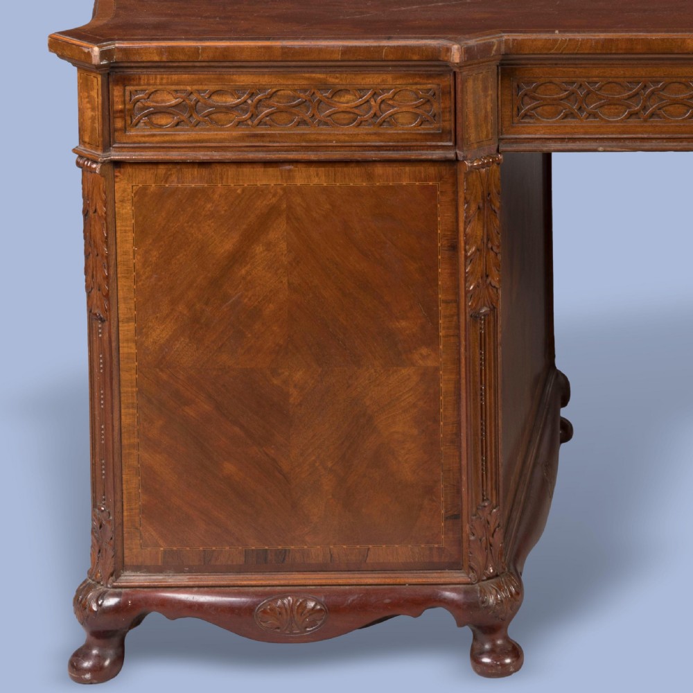 An antique mahogany pedestal desk featuring a serpentine front, parquetry drawer fronts, brass handles, and carved blind fretwork along the frieze. View VI