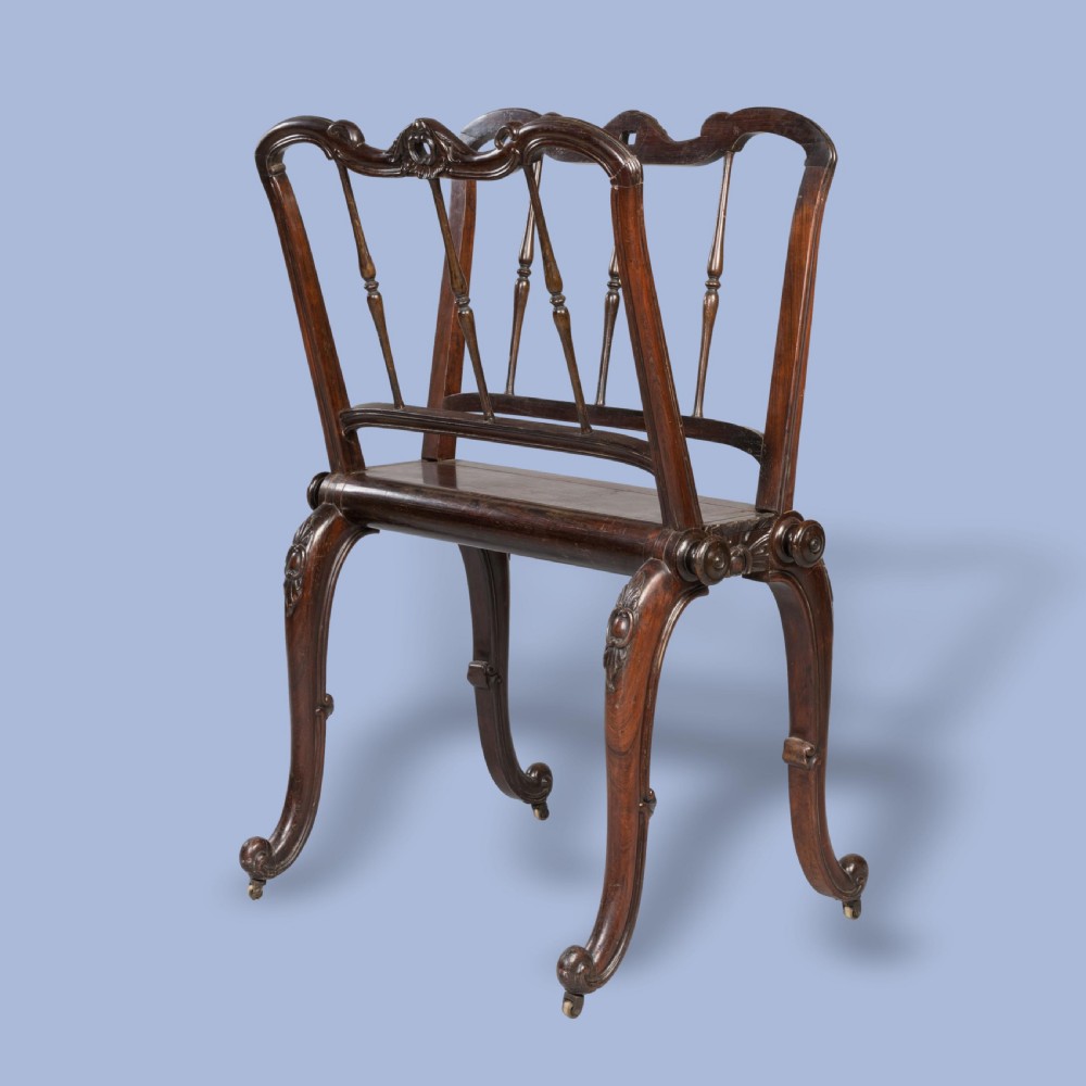 A late Georgian dark mahogany folio stand featuring carved slatted side supports, turned spindles, curved legs, and small brass castors.