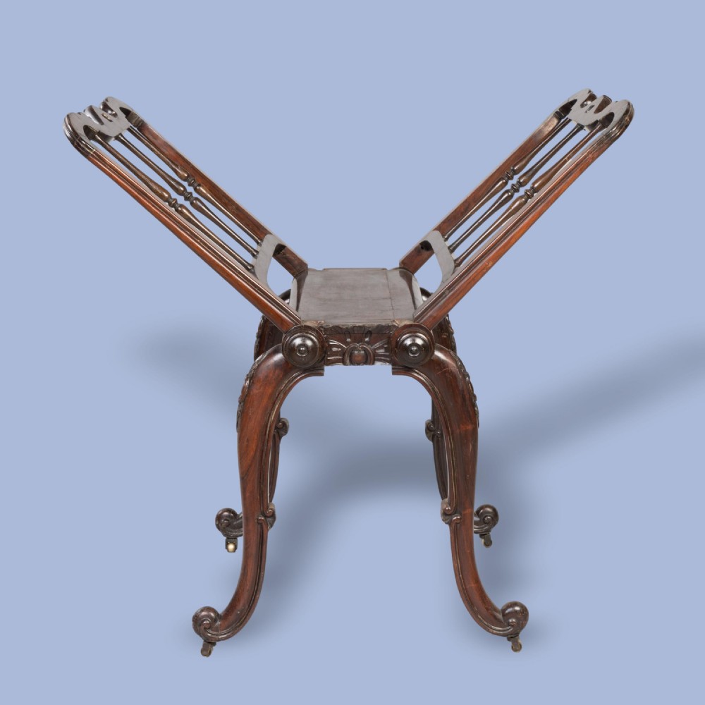A late Georgian dark mahogany folio stand featuring carved slatted side supports, turned spindles, curved legs, and small brass castors. View III