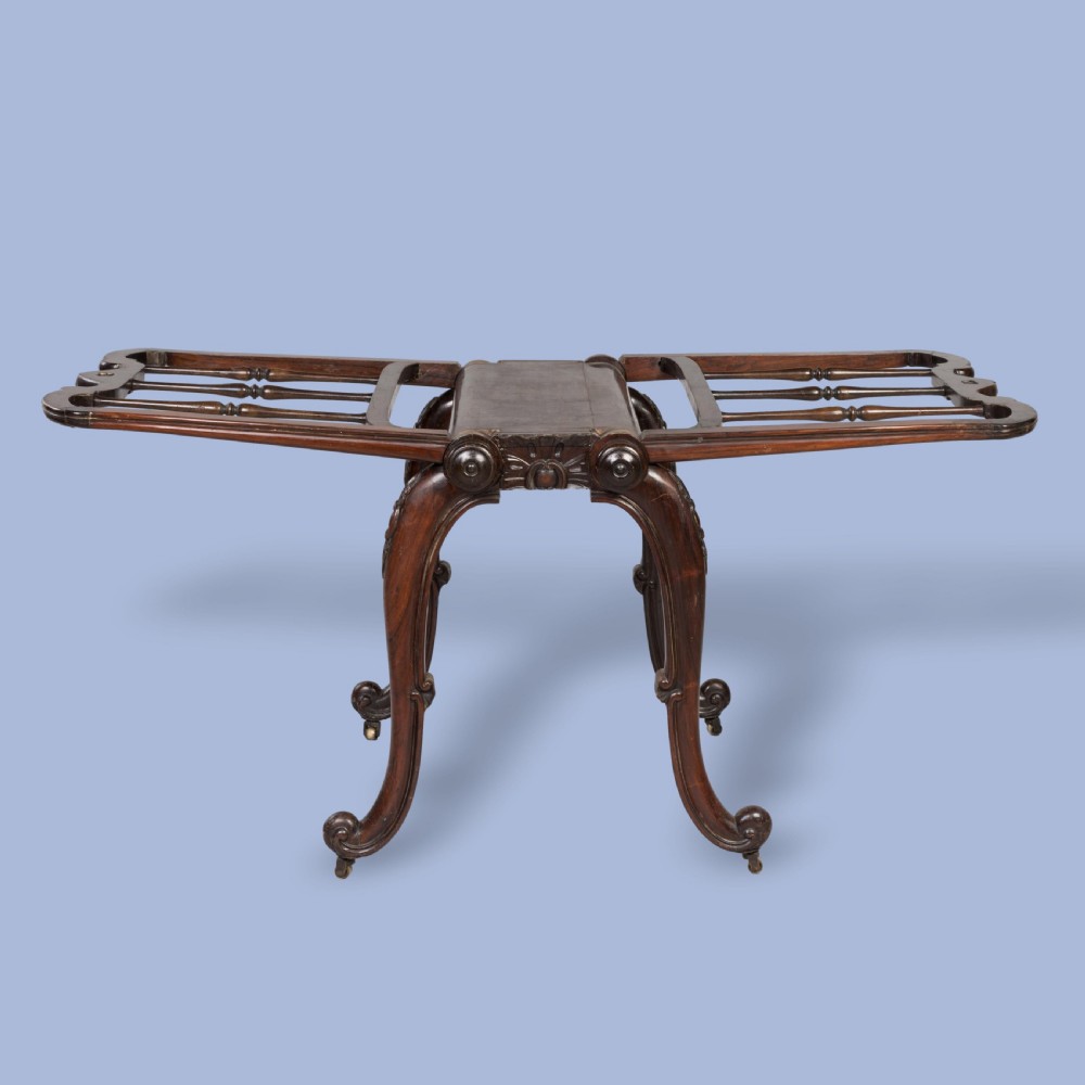 A late Georgian dark mahogany folio stand featuring carved slatted side supports, turned spindles, curved legs, and small brass castors. View IV