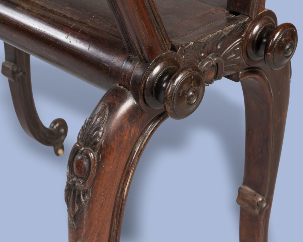 A late Georgian dark mahogany folio stand featuring carved slatted side supports, turned spindles, curved legs, and small brass castors. View V