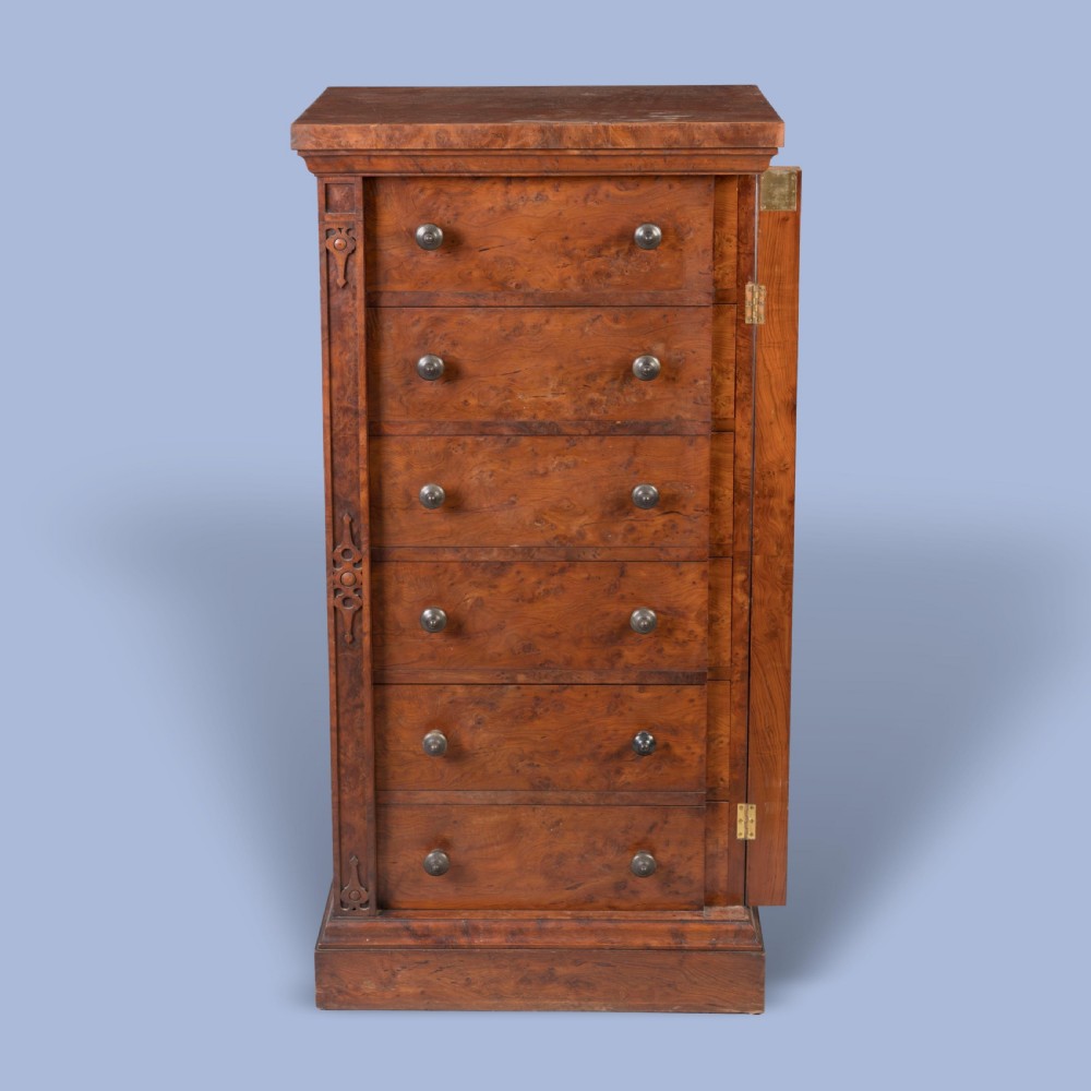 A tall, narrow Victorian Wellington chest of drawers featuring six drawers crafted from highly figured warm brown timber with carved side stiles and turned wooden knob handles. View VI
