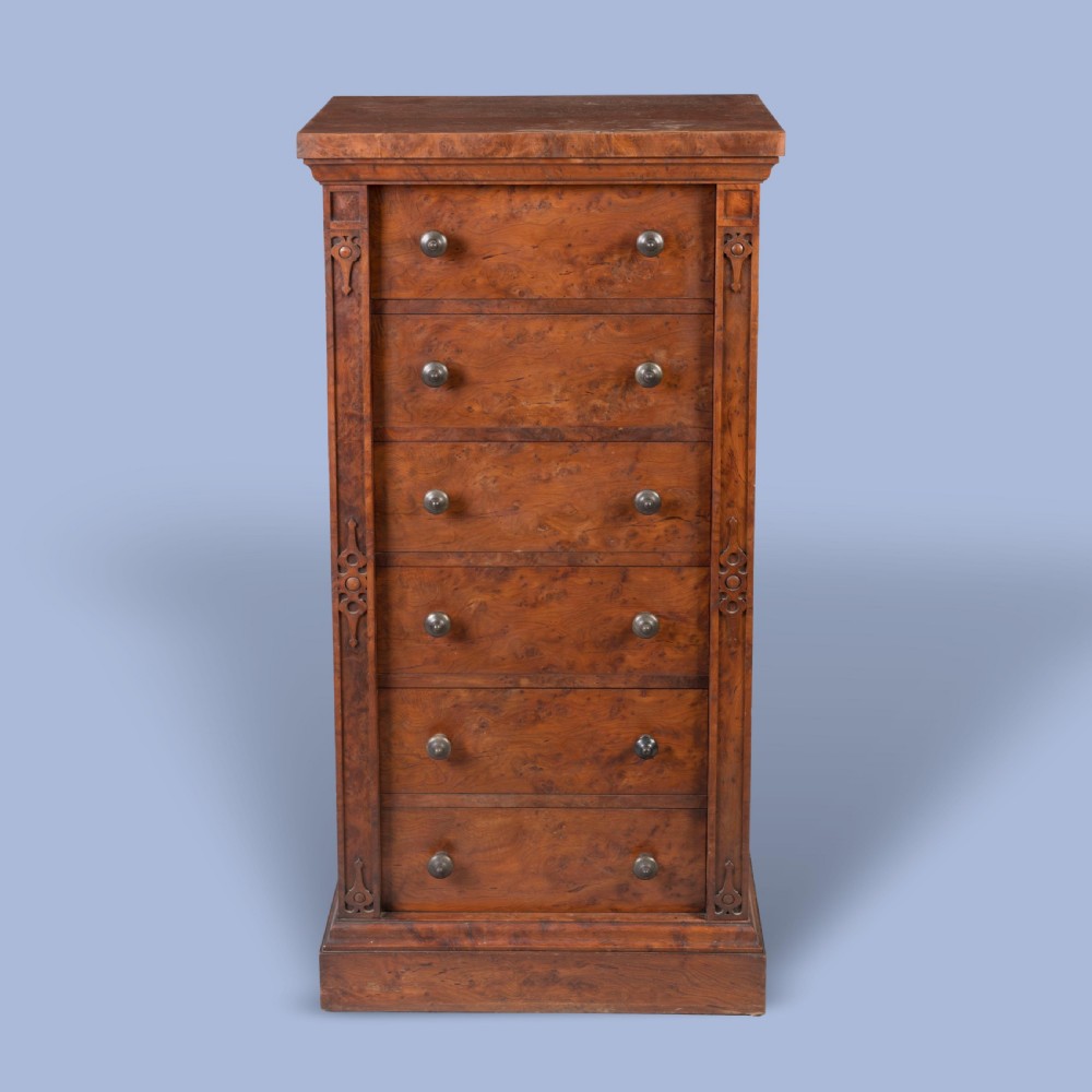 A tall, narrow Victorian Wellington chest of drawers featuring six drawers crafted from highly figured warm brown timber with carved side stiles and turned wooden knob handles.