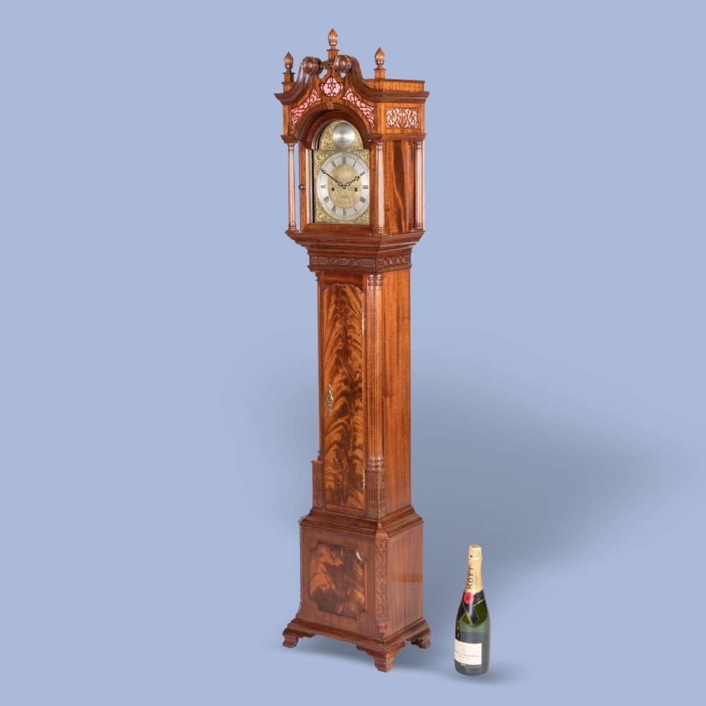 A miniature mahogany longcase clock featuring a brass dial, swan-neck pediment with brass finials, and a flame mahogany veneer trunk door. View VI