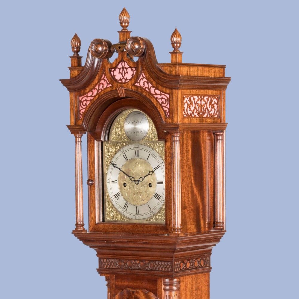 A miniature mahogany longcase clock featuring a brass dial, swan-neck pediment with brass finials, and a flame mahogany veneer trunk door. View IV