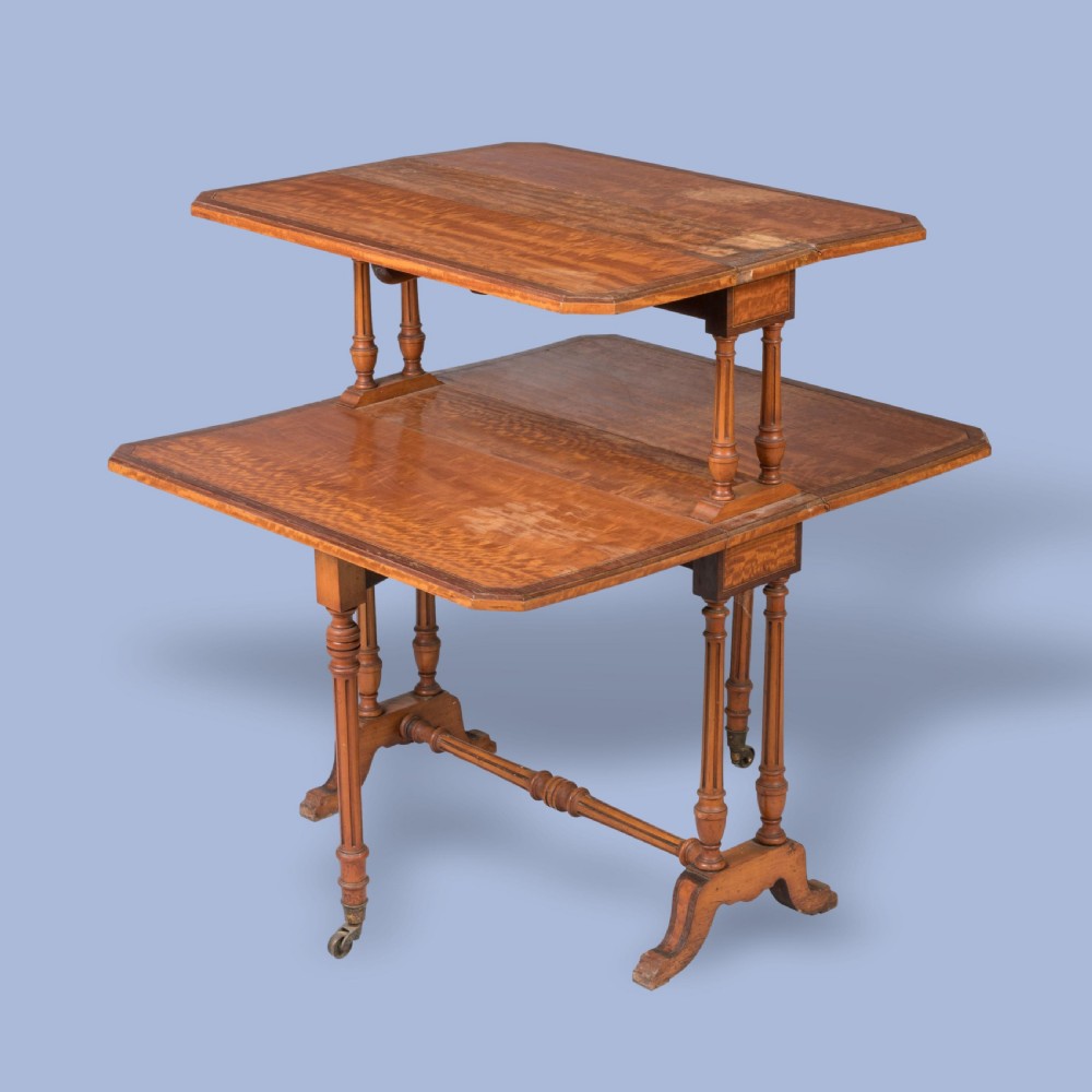 A double-tiered satinwood Sutherland drop-leaf table featuring turned leg supports, crossbanded borders, and brass castors.