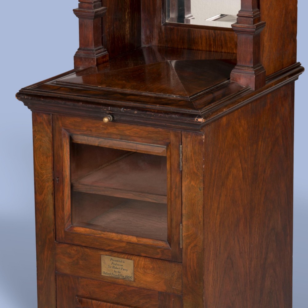 A dark mahogany Victorian music cabinet featuring a pedimented top, an upper section with open horizontal shelves, a glazed lower door, a brass presentation plaque, and small castors at the base. View II
