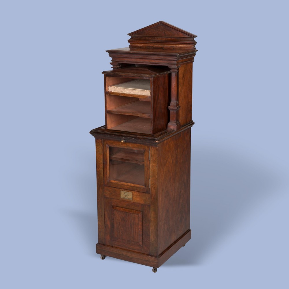 A dark mahogany Victorian music cabinet featuring a pedimented top, an upper section with open horizontal shelves, a glazed lower door, a brass presentation plaque, and small castors at the base.