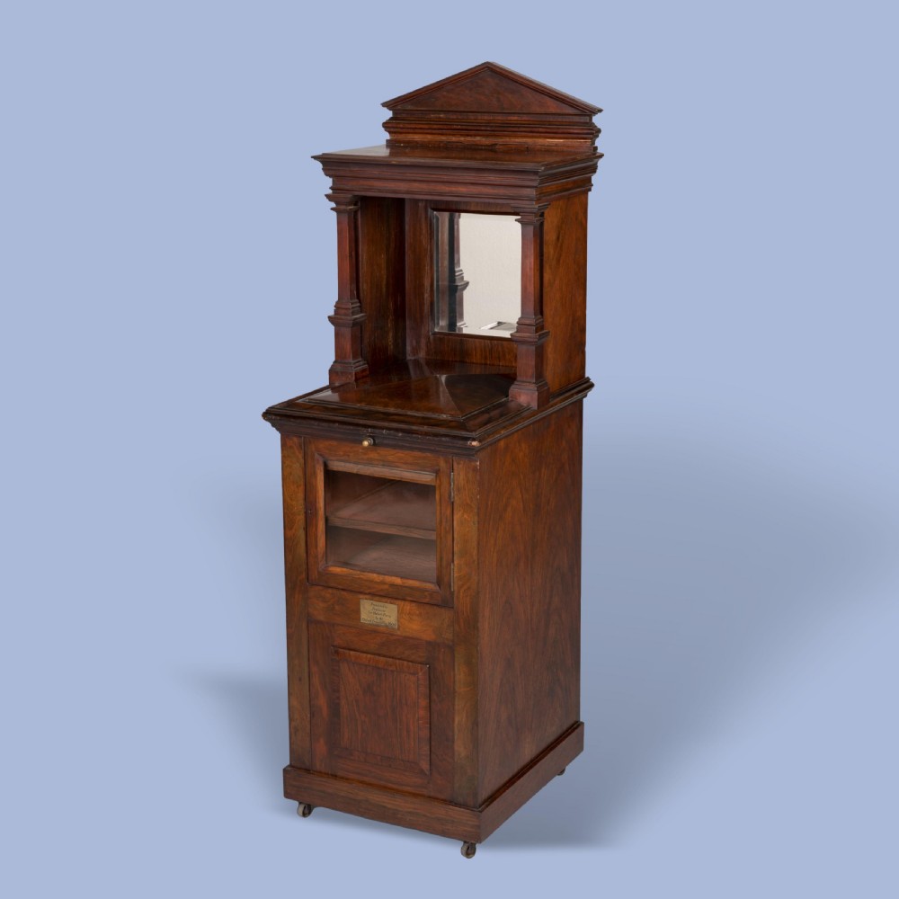 A dark mahogany Victorian music cabinet featuring a pedimented top, an upper section with open horizontal shelves, a glazed lower door, a brass presentation plaque, and small castors at the base. View VI
