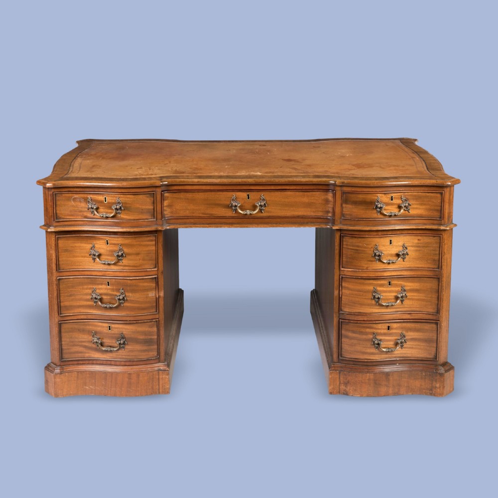 A George III style mahogany pedestal library desk featuring a leather writing surface, serpentine contoured front, nine drawers, and brass bail handles.