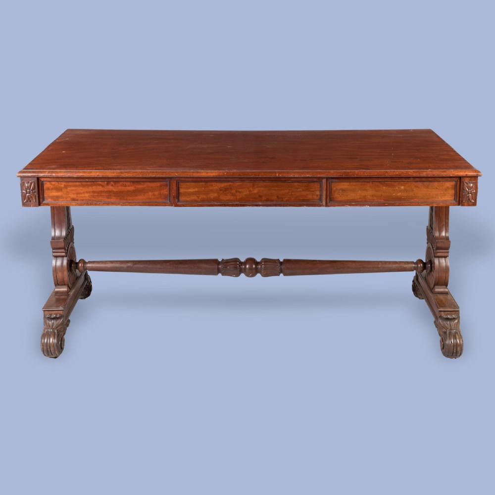 A Regency mahogany library table featuring a flat rectangular top, three frieze drawers, carved trestle end supports, and a central turned stretcher.