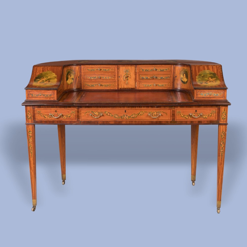 A hand-painted satinwood Carlton House desk featuring a curved superstructure of drawers, classical portrait medallions, floral swags, a leather writing surface, and square tapered legs on castors.